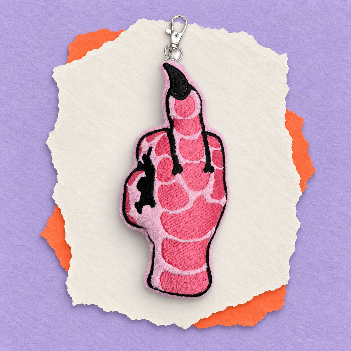 A pink embroidered pigeon foot keychain charm showing the middle finger, with black fingernails and black outlines, attached to a silver lobster clasp. The charm is displayed on a layered background of torn white paper and orange paper against a solid purple background.